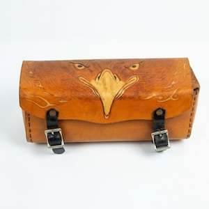 May include: A tan leather cylindrical bag featuring a detailed embossed eagle face on the front flap. The bag is secured with two black leather straps that have silver-toned metal buckles. The edges are stitched with dark brown thread.