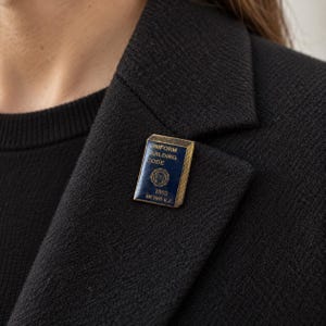 May include: A rectangular metal lapel pin featuring a dark blue enamel finish with gold-toned edges. The pin displays the text UNIFORM BUILDING CODE, the year 1993, and METRO K.C. along with a circular emblem in the center of the book-shaped design.