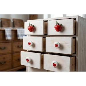 May include: A light-colored wooden chest of drawers featuring six drawers. The top two drawers have three-dimensional red apple-shaped knobs with green leaves. The middle and bottom drawers have round white knobs with a small red apple graphic.