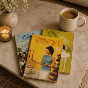 May include: A collection of vintage magazines, a lit candle in a glass holder, a coffee cup, and a small vase of white flowers. The magazines have yellow, blue, and green covers with the text "The Workbasket" and "The Work and Home Arts Magazine".