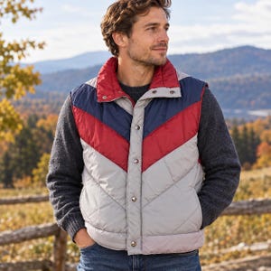 May include: A quilted puffer vest featuring a chevron color-block pattern in navy blue, red, and light gray. The vest has a high collar, a front snap-button closure, and is worn over a dark gray long-sleeve sweater and blue denim jeans.