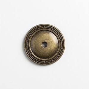 May include: A circular cabinet knob with an antique brass finish. The knob features a smooth, slightly domed center and a raised outer border decorated with a repeating geometric Greek key pattern. A single screw hole is centered in the middle.