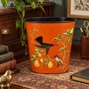 May include: A cylindrical metal wastebasket with a vibrant orange, hammered texture finish. The exterior features a detailed illustration of two birds perched on branches with yellow flowers and green leaves. The rim is a solid black band.