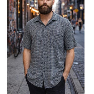 May include: Short-sleeved button-down shirt with a small, checkered pattern in shades of gray and white. The shirt has a classic collar and a single chest pocket. The person is wearing black pants.