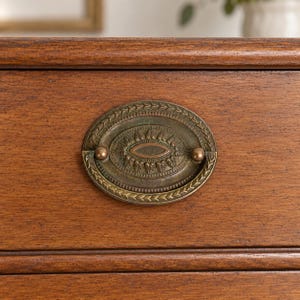 May include: An oval-shaped brass drawer pull with an intricate embossed border and a central eye-like motif, mounted on a smooth, medium-brown wooden surface. The metal hardware features a dark, aged patina with visible texture and detail.