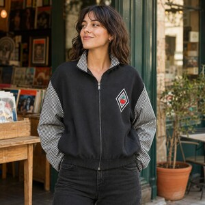 May include: A black zip-up jacket featuring long sleeves with a black and white gingham pattern, a matching gingham collar, and a diamond-shaped patch on the chest containing a small red apple graphic. The jacket has a ribbed hem and cuffs.