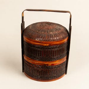 May include: A cylindrical, two-tiered woven basket with a dark brown finish. The basket features a domed lid and a tall, arched handle that extends from the base to the top. The structure is composed of tightly woven strips of material.