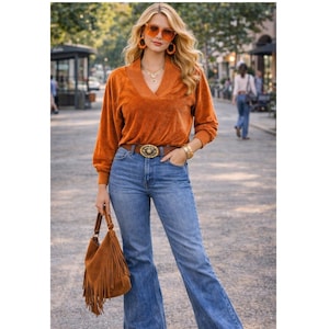 Puede incluir: Una mujer con un top de terciopelo naranja, vaqueros azules acampanados y un cinturón marrón con hebilla decorativa. Lleva un bolso de flecos de ante marrón y gafas de sol y pendientes naranjas. El conjunto tiene un estilo retro y bohemio.