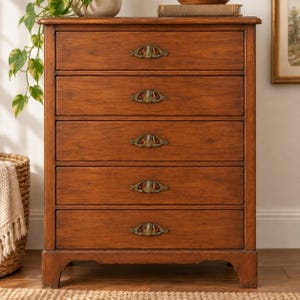 Puede incluir: Una cómoda alta de madera con cinco cajones horizontales, cada uno equipado con un tirador decorativo de metal ornamentado. El mueble tiene un acabado en tono madera marrón medio cálido con veta visible y se apoya sobre cuatro patas.