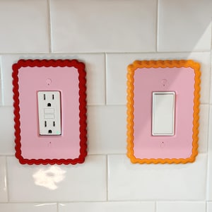 May include: Two rectangular wall plates with scalloped edges. The left plate is red with a pink center and a white electrical outlet. The right plate is orange with a pink center and a white light switch. Both are mounted on a white tiled wall.