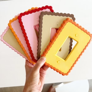 May include: A collection of rectangular light switch plates with scalloped edges, arranged in a fan shape. The plates feature a variety of solid colors including yellow, orange, red, pink, and brown, each with two small circular screw holes.