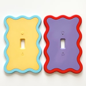 May include: Two rectangular light switch plates with wavy, scalloped edges. The left plate is yellow with a light blue border. The right plate is purple with a red border. Each plate features a central rectangular cutout and two screw holes.