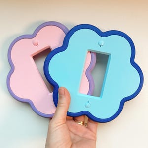 May include: Two flower-shaped light switch plates. One plate is light pink with a purple border, and the other is light blue with a dark blue border. Each plate features a rectangular center cutout and two small circular screw holes.