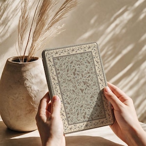 May include: A rectangular e-reader device featuring a sage green cover with a dense, intricate pattern of small brown leaves and tiny white flowers, framed by a decorative border of similar floral motifs in muted earth tones.