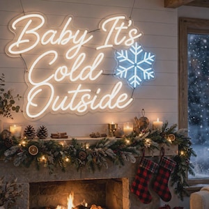 Op de afbeelding: Een wit neonbord met de tekst Baby It's Cold Outside in een cursief lettertype, vergezeld van een lichtblauw neon sneeuwvlokpictogram. Het bord is gemonteerd op een witte wand met horizontale planken boven een schoorsteenmantel.
