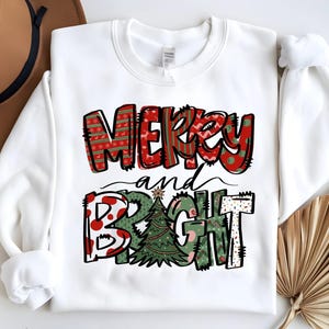 May include: A white long-sleeve sweatshirt featuring the text Merry and Bright in a stylized font. The word Merry is in red with green patterns, and the word Bright is in green with a Christmas tree replacing the letter I.