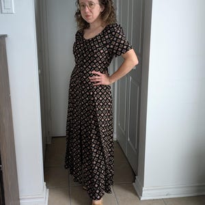 May include: A woman wearing a black maxi dress with a geometric pattern and brown loafers. The dress has a scoop neck and short sleeves.