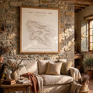 Può includere: Una stampa di mappa topografica incorniciata intitolata FIELD I - SANTA LUCIA - 280M, con linee di contorno marroni su sfondo crema, in una cornice di legno chiaro su una parete in pietra sopra un divano beige con cuscini e una coperta.