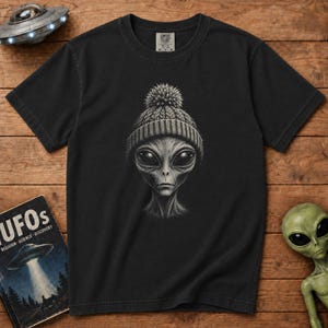 Könnte beinhalten: Ein schwarzes Kurzarm-T-Shirt mit einer Grafik eines Außerirdischen, der eine Strickmütze mit Bommel trägt. Das T-Shirt hat einen Rundhalsausschnitt und ein Markenetikett von Comfort Colors am inneren Halsausschnitt.