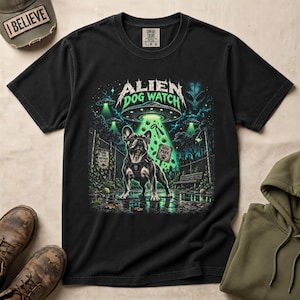 May include: A black t-shirt featuring a graphic print of a French Bulldog wearing a harness in a park at night. Above the dog, a UFO emits a green beam of light. The text reads ALIEN DOG WATCH, with smaller signs saying DOG PARK and LANE YARD DIFFERENT GALAXY.
