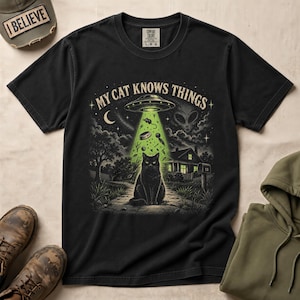 May include: A black short-sleeve t-shirt featuring a graphic print of a black cat sitting under a glowing green UFO beam. The text MY CAT KNOWS THINGS is printed above the UFO. The design includes a house, a crescent moon, and an alien face.