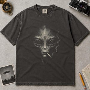 Könnte beinhalten: Ein anthrazitfarbenes Kurzarm-T-Shirt mit einer detaillierten Grafik eines Alien-Gesichts mit großen dunklen Augen und einer Zigarette im Mund, aus der ein dünner Rauchfaden aufsteigt. Das T-Shirt hat einen Rundhalsausschnitt und ein Etikett.