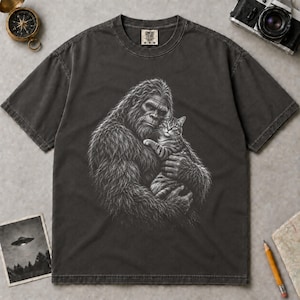 May include: A charcoal gray short-sleeve t-shirt featuring a white graphic print of a large, hairy creature holding a tabby cat. The shirt has a crew neckline and a Comfort Colors brand label at the inner neck. The fabric has a washed, vintage look.