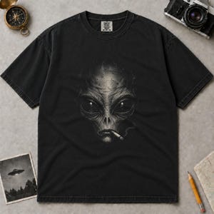 May include: A black short-sleeved t-shirt featuring a graphic print of an alien face with large dark eyes and a cigarette in its mouth. The shirt has a crew neckline and a Comfort Colors brand label at the inner collar. The fabric has a washed-out look.