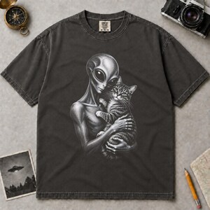 May include: A charcoal gray short-sleeve t-shirt featuring a detailed black and white graphic print of an alien holding a tabby cat. The shirt has a crew neckline and a label at the inner collar that reads Comfort Colors with the size M.