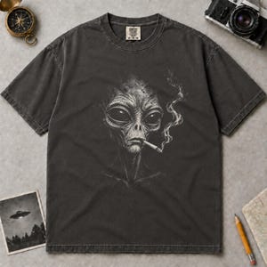 Könnte beinhalten: Ein anthrazitfarbenes Kurzarm-T-Shirt mit einer weißen grafischen Illustration eines Aliens, das eine Zigarette raucht. Das T-Shirt hat einen Rundhalsausschnitt und ein Comfort Colors Markenetikett an der Innenseite des Kragens.