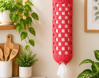 May include: A cylindrical, coral-colored crochet bag designed for storage, featuring an open-weave grid pattern. The bag hangs from a wooden peg on the wall and contains white plastic bags, with one bag protruding from the bottom opening.
