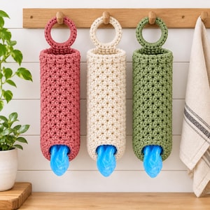 May include: Three cylindrical crochet bag dispensers in dusty rose, cream, and sage green. Each features a circular top loop for hanging and a star-stitch pattern. A bright blue plastic bag is visible protruding from the bottom opening of each.
