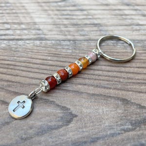 May include: A beaded keychain with a silver key ring and a pendant. The keychain features a series of colorful beads in shades of red, orange, and pink. The pendant is a silver oval with a cross.