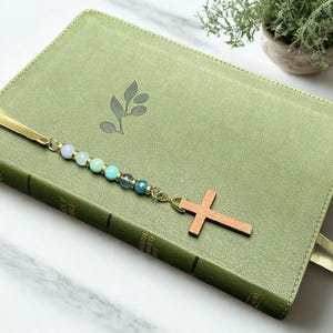 May include: A sage green book with a gold ribbon bookmark attached to a beaded chain. The chain features six round beads in shades of light blue, turquoise, and dark blue, ending with a simple wooden cross charm. A small leaf icon is centered.
