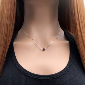 May include: A delicate silver chain necklace featuring a single, small, faceted deep purple gemstone bead centered on the strand. The necklace rests against the neck, showcasing a minimalist design with a thin metallic chain and a dark violet stone.