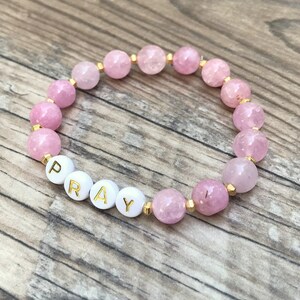 May include: A beaded bracelet featuring round, faceted light pink gemstone beads interspersed with small gold-toned metal spacers. Four white circular beads with gold-colored lettering spell out the word PRAY, integrated into the bead sequence.