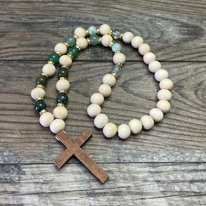 May include: A rosary with wooden beads and a wooden cross. The beads alternate between light wood and green stone, connected by gold-colored spacers. The cross is a dark brown color.