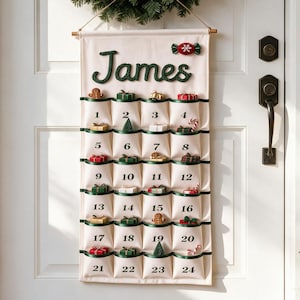 Può includere: Un calendario dell'avvento in tessuto color crema appeso a una porta, con il nome James in corsivo verde scuro, un ornamento a forma di caramella rossa e ventiquattro tasche numerate da uno a ventiquattro, contenenti piccoli oggetti natalizi.