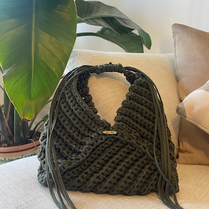 Può includere: Una borsa a mano all'uncinetto color verde oliva scuro con silhouette triangolare, un manico integrato e lunghe frange di corda pendenti su entrambi i lati. La borsa è chiusa al centro da una piccola chiusura a levetta color oro.