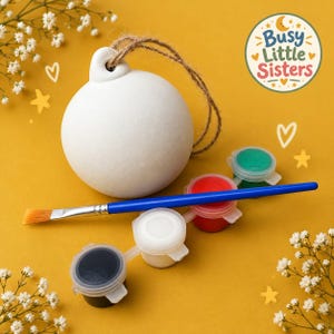 May include: A white ceramic ornament sphere with a jute twine loop, accompanied by a blue paintbrush and four small plastic paint pots containing black, white, red, and green paint. A circular logo reads Busy Little Sisters.
