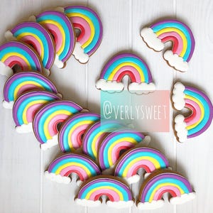 May include: A collection of rainbow-shaped cookies decorated with colorful icing in shades of purple, blue, yellow, and pink, featuring white clouds at each end. A semi-transparent rectangular overlay displays the text @VERLYSWEET in white.