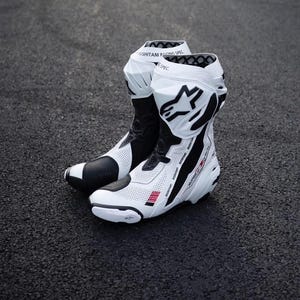 Puede incluir: Un par de botas de carreras de moto blancas y negras con paneles de cuero perforado, un talón reforzado y un logotipo de Alpinestars prominente en el lateral. El puño superior muestra el texto Kushitani Racing Spec en letras negras.
