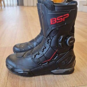 May include: A pair of black motorcycle boots featuring a high-top design with red BSP branding on the upper shaft. The boots include a dial-based lacing system on the side, reinforced toe and heel sections, and textured panels for added durability.