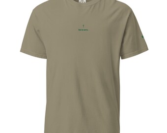 May include: A khaki-colored short-sleeve t-shirt featuring a small green cross icon centered on the chest above the text "but to serve." The shirt has a crew neckline and a small green graphic detail visible on the edge of the left sleeve.