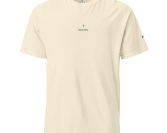 May include: A cream-colored short-sleeve t-shirt featuring a small green cross icon above the text "but to serve." centered on the chest. The sleeve displays a partial green graphic. The shirt has a crew neckline and a standard fit design.