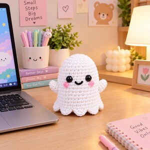 May include: A white crocheted ghost plushie with black plastic eyes, a stitched black mouth, and pink crocheted cheeks. The ghost has a rounded head, small arms extending to the sides, and a scalloped bottom edge, sitting on a wooden desk surface.