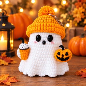 May include: A white crocheted ghost keychain wearing an orange beanie with a pom-pom. The ghost has black bead eyes, pink cheeks, and a small black mouth. It holds a small orange coffee cup in one hand and a tiny orange jack-o'-lantern in the other.