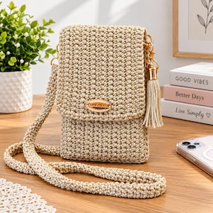 Op de afbeelding: Een beige gehaakte schoudertas met een lange riem en een overslagklep. De tas is voorzien van een ovaal goudkleurig metalen label met het woord handmade gegraveerd op de voorzijde en een bijpassend beige kwastje aan een goudkleurige sluiting.