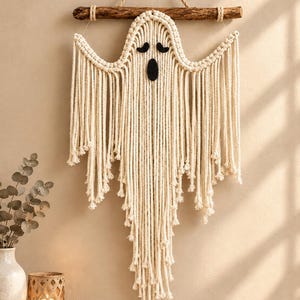 May include: A macrame wall hanging shaped like a ghost, crafted from off-white cotton cord. The ghost features two black felt eyes and an oval black felt mouth. It is suspended from a natural wooden branch with a thick jute rope hanger.