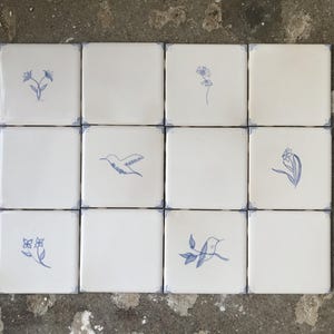 Puede incluir: Un conjunto de doce azulejos de cerámica cuadrados dispuestos en cuadrícula. Cada azulejo es de color blanco roto con acabado brillante y presenta un delicado dibujo de líneas azules. Los diseños incluyen motivos florales y colibríes con esquinas azules.